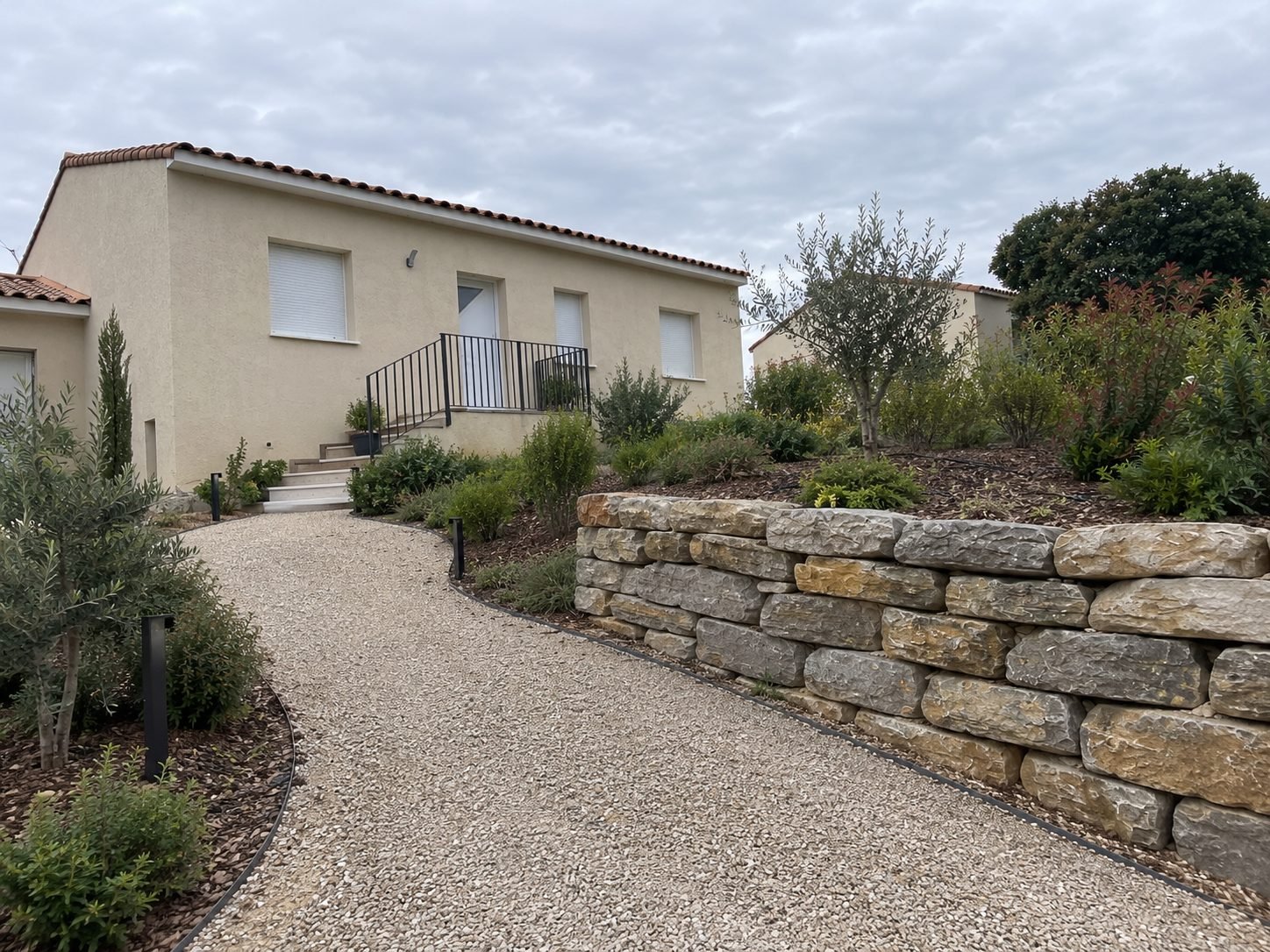 Allée en gravier bordée de massifs plantés et d'un muret en pierres sèches, jusqu'à l'entrée de la maison