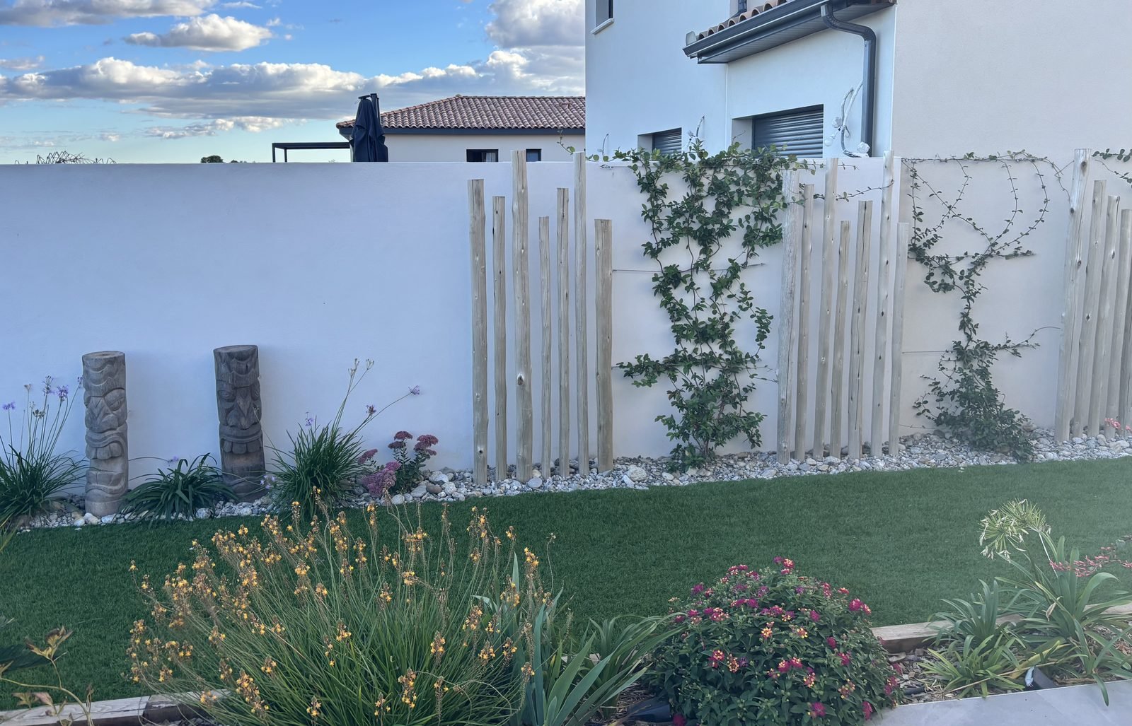 Massif fleuri avec piquets en bois et plantes grimpantes le long d'un mur blanc, devant le gazon